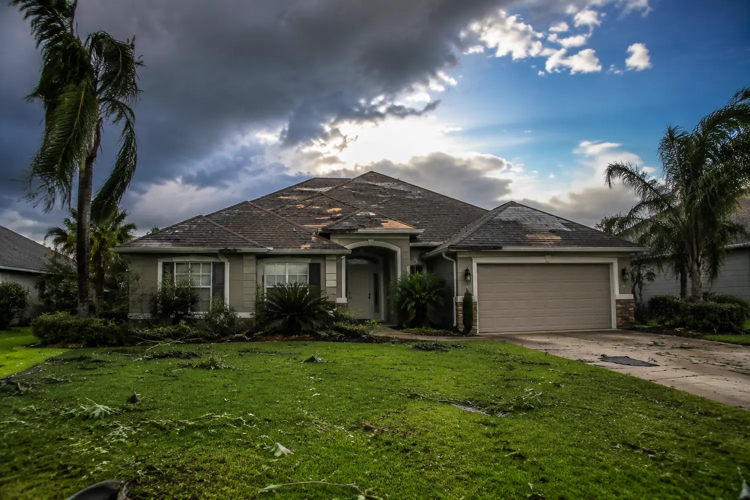 A Florida home with visible roof shingle damage after a storm, debris in the yard, blue sky emerging